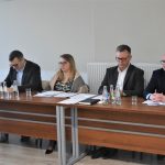 Trzech dorosłych mężczyzn w garniturach i kobieta zasiadających za długim biurkiem.