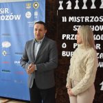 Młody mężczyzna w garniturze i kobieta w jasnym swetrze stojący przed ścianką.