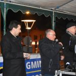 Trzech mężczyzn ubranych w ciemne płaszcze przemawiają na scenie do zgromadzonych.