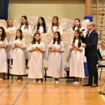 Dorośli mężczyzna i kobieta oraz grupa dziewcząt przebranych w strój aniołów. Wszyscy znajdują się na sali gimnastycznej.