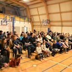 Duża grupa osób różnej płci i wieku siedząca na sali gimnastycznej.