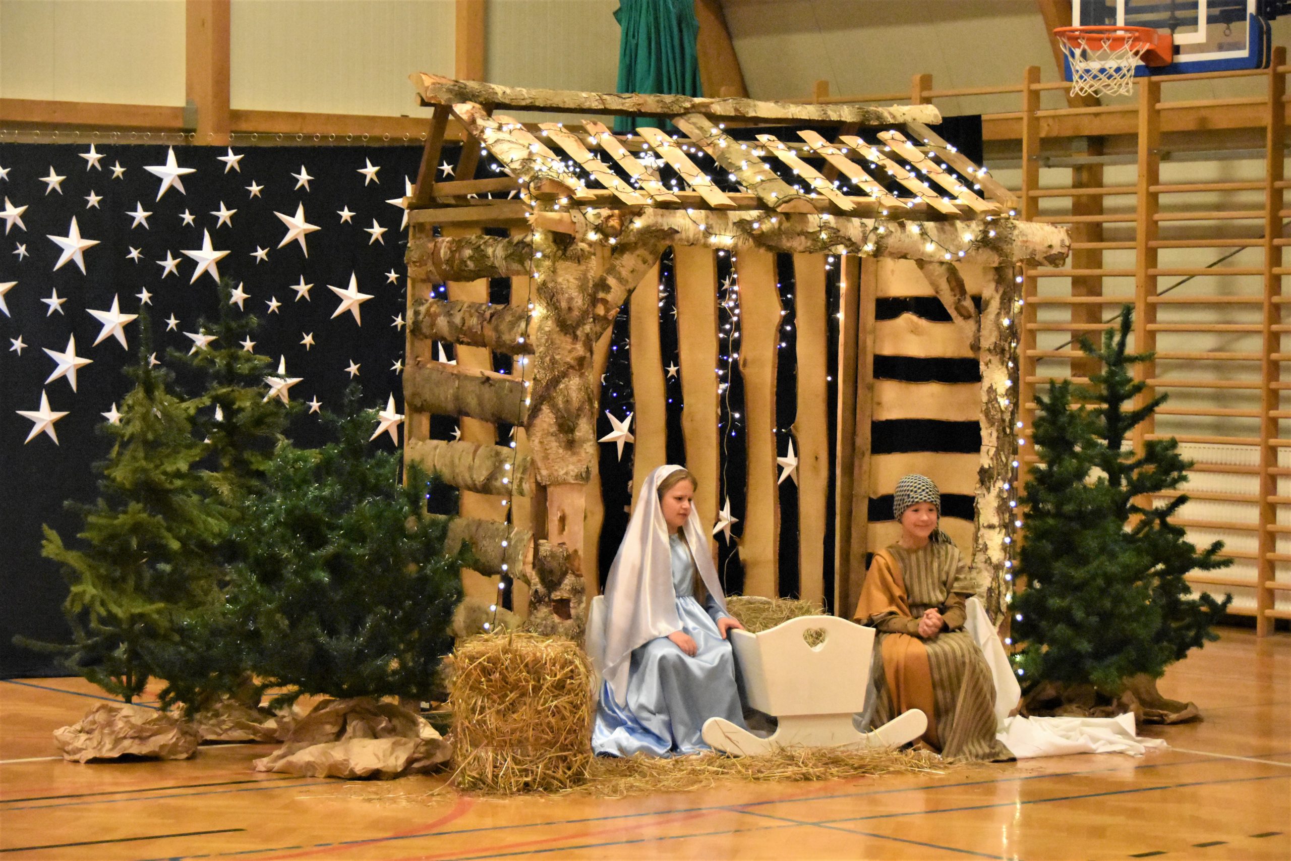 Drewniana szopka na sali gimnastycznej. Siedzi w niej dwójka dzieci przebranych za Maryję i Józefa.