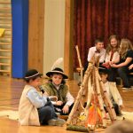 Grupa dzieci siedząca na sali gimnastycznej. Część z nich ubrana w góralskie stroje siedzi przy ozdobie pozorującej ognisko.