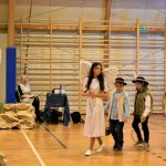 Dziewczyna w stroju anioła idąca przez sale gimnastyczną. Za nią podąża trójka chłopców w stroju góralskim.