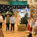 Grupa dzieci w strojach jasełkowych odgrywająca scenę teatralna przed drewnianą szopką na sali gimnastycznej.