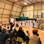 Grupa osób różnej płci i wieku na sali gimnastycznej podziwia występ jasełkowy.