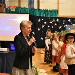 Starsza kobieta przemawia na sali gimnastycznej. Obok niej stoją dzieci ubrane w jasełkowe stroje.