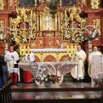 Mężczyźni ubrani w szaty stoją w kościele przy ołtarzu, koloru czerwono-złotego.