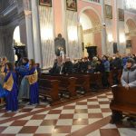 Grupa osób siedzi w ławkach w kościele, z boku stoi choinka, i barwny korowód.