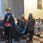 Młody chłopak przebrany w długą szatę, i zieloną czapkę z koroną stoi w kosciele. Za nimi dziewczyny stoją i trzymają mu płaszcz.