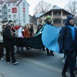 Grupa osób idzie w pfrzemarszu po ulicy. Młode dziewczyny niosą szatę, za nimi idą starsze osoby ubrane w czerwone szaty.