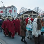 Grupa osób idzie w pfrzemarszu po ulicy. Młode dziewczyny niosą szatę, za nimi idą starsze osoby ubrane w czerwone szaty.
