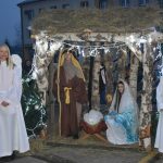 Szopka z drewana piękne oświetlona. W niej siedzi dziewczyna ubrana w niebieską szatę, trzyma w rękach małe dzieciątko ubrane na biało, obok stoi mężczyzna ub rany w sztae brązową. Obok szopki stoją dziewczyny ubrane w białe sukienki ze skrzydłami