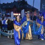 Mężczyzna przebrany w długą szatę niebieską, obok niego dziewczyny niosące jego płaszcz, idzie w przemarszu po koscte brukowej,