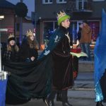 Młody chłopak przebrany w długi płaszcz niesie w rękach krzynkę, za nimi dziewczyny trzymające jego płaszcz.