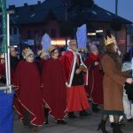 Starszy mężczyzna ubrany w czerwoną szatę obok niego starsze kobiety ubrane na czerwono idą w przemarszu.