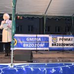 Młoda kobieta w płaszczu jasnhym i jasnych włosahc przemawia do mikrofonu na scenie.