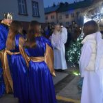 Trzy osoby ubrane w niebieskie szaty, składają dary do szopki. Obok szopki stoją dziewczyny ubrane na biało.