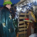 Młody mężczyzna ubrany w długą szatę zieloną składa dary do szopki.