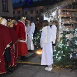 Grupa osób ubranych na czerwono składa dary do szopki.