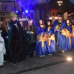 Grupa osób ubranych w niebieskie i zielone szaty stoją przed sceną na rynku.