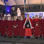 Grupa osób ubranych w czerwone szaty stoją na rynku przedsceną.