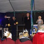 Trzech mężczyzn stoi n a scenie. Jeden z nich trzyma w rękach mikrofn i przemawia.