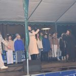 Grupa dzieci spiewa na scenie. Na czele kobieta ubrana w jasny płaszcz.