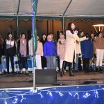 Grupa dzieci spiewa na scenie. Na czele kobieta ubrana w jasny płaszcz.