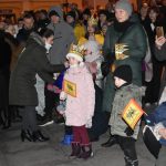 Grupa osób stoi na rynku podczas wydarzenia imprezowego. Młoda kobieta wręcza dzieciom cukierki.