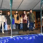 Grupa dziewczyn i chłopców spiwa na scenie. W trękach trzymają mikrofny,