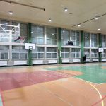 Widok na rozległą salę gimnastyczną. Na jednej ze ścian znajdują sie okna. Stoi przy nich dwóch mężczyzn.