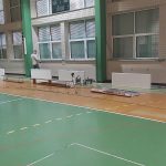 Widok na rozległą salę gimnastyczną. Na jednej ze ścian znajdują sie okna pokryte siatką. Stoi przy nich meżczyzna..