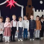 Grupa dzieci w czasie przedatwienia jasełkowego. Stoją na ozdobnym podescie i pozują w rzędzie do zdjęcia.