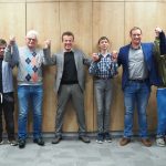 Grupa mężczyzn stojąca przed ścianą i trzymajaca się za ręce.