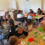 Grupa dzieci na rozległej sali w czasie warsztatów plastycznych.