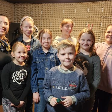 Grupa dzieci pozuje do zdjęcia z młodą kobietą w studiu nagłośnieniowym.