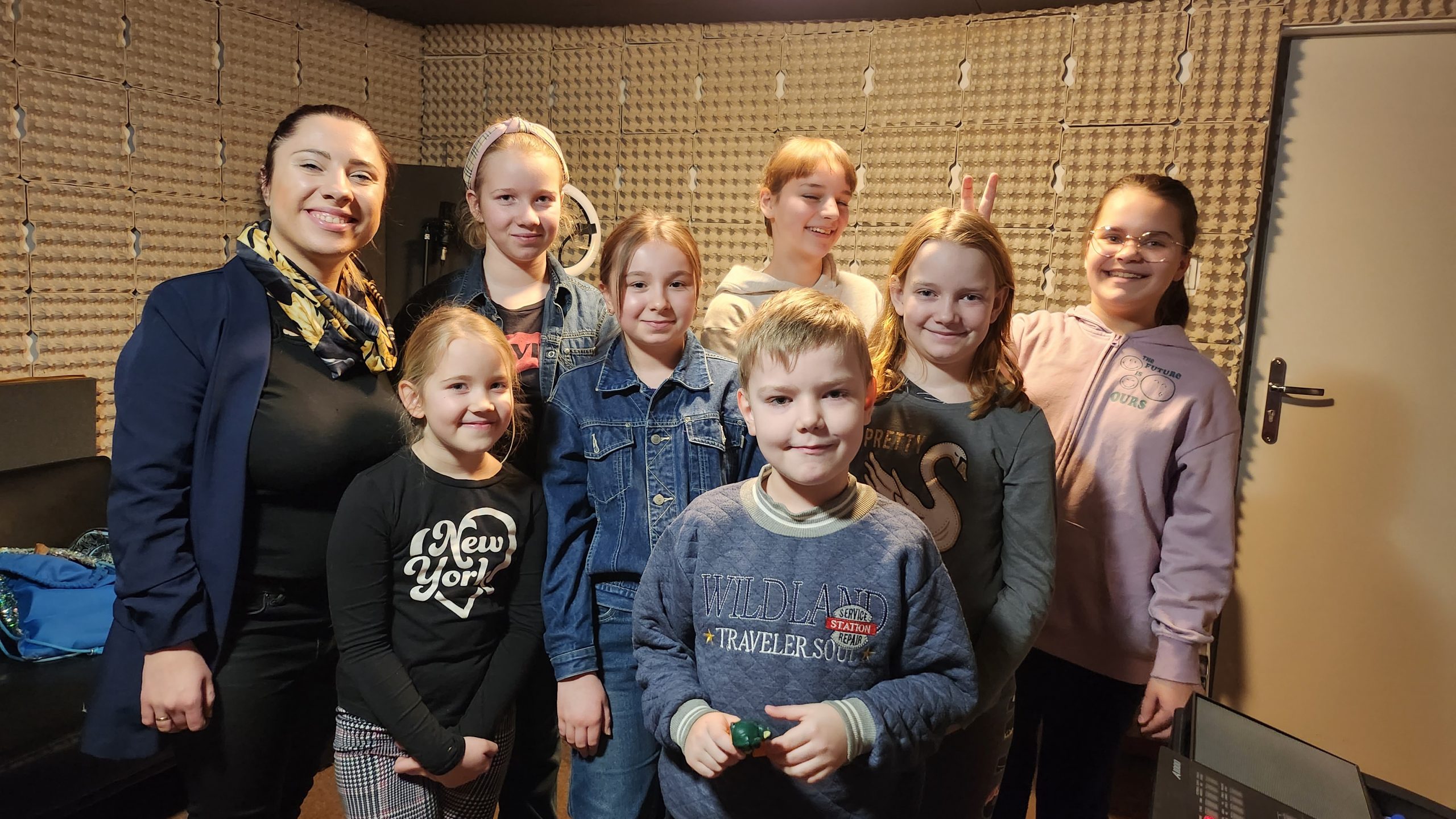 Grupa dzieci pozuje do zdjęcia z młodą kobietą w studiu nagłośnieniowym.