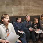 Grupa dzieci siedzi w sali i uczy się spiewu. Trzymają w rękach kartki.