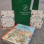 Torba prezentowa koloru zielonego, dyplomy oraz książka w twardej, kolorowej oprawie leżą na dywanie.