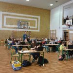 Grupa dzieci siedzi na sali przy stolikach, na których znajdują sie laptop, na których programują.