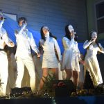 Młode dziewczyny i chłopcy ubrani na biało spiewają do mikrofonu na scenie.