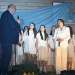 Starszy mężczyzna w garniturze przeamawia do scenie do kobiety ubranej w biały kombinezon. Za nimi młode dziewczyny ubrane na biało.