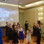 Grupa dzieci ubranych w odświetne stroje śpiewają do mikrofonu na sali.