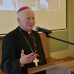 Starszy mężczyzna ubrany w sutannę przemawia do mikrofonu. Na szyi wisi mu krzyż.