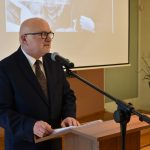 Starszy mężczyzna w garniturze i w okularach przemawia do mikrofonu na konferencji.