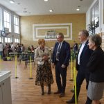 Grupa starszych osób stoi na sali i patrzy na rywalizację dzieci podczas konkurncji poruszania samochodów zbudowanych z klocków lego.