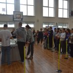 Grupa dzieci stoi podczas zawodów i patrzy jak kilka osób obsługiek samochody zbudowane z klocków lego.