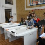 Grupa dzieci stoją przy stołach i bawią się samoochodzikacmi zbudowanymi z klocków lego.