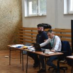 Dwóch mężczyzn siedzi przy stoliku na którym stoi laptop. Obok nich stoi duży monitor z wynikami.